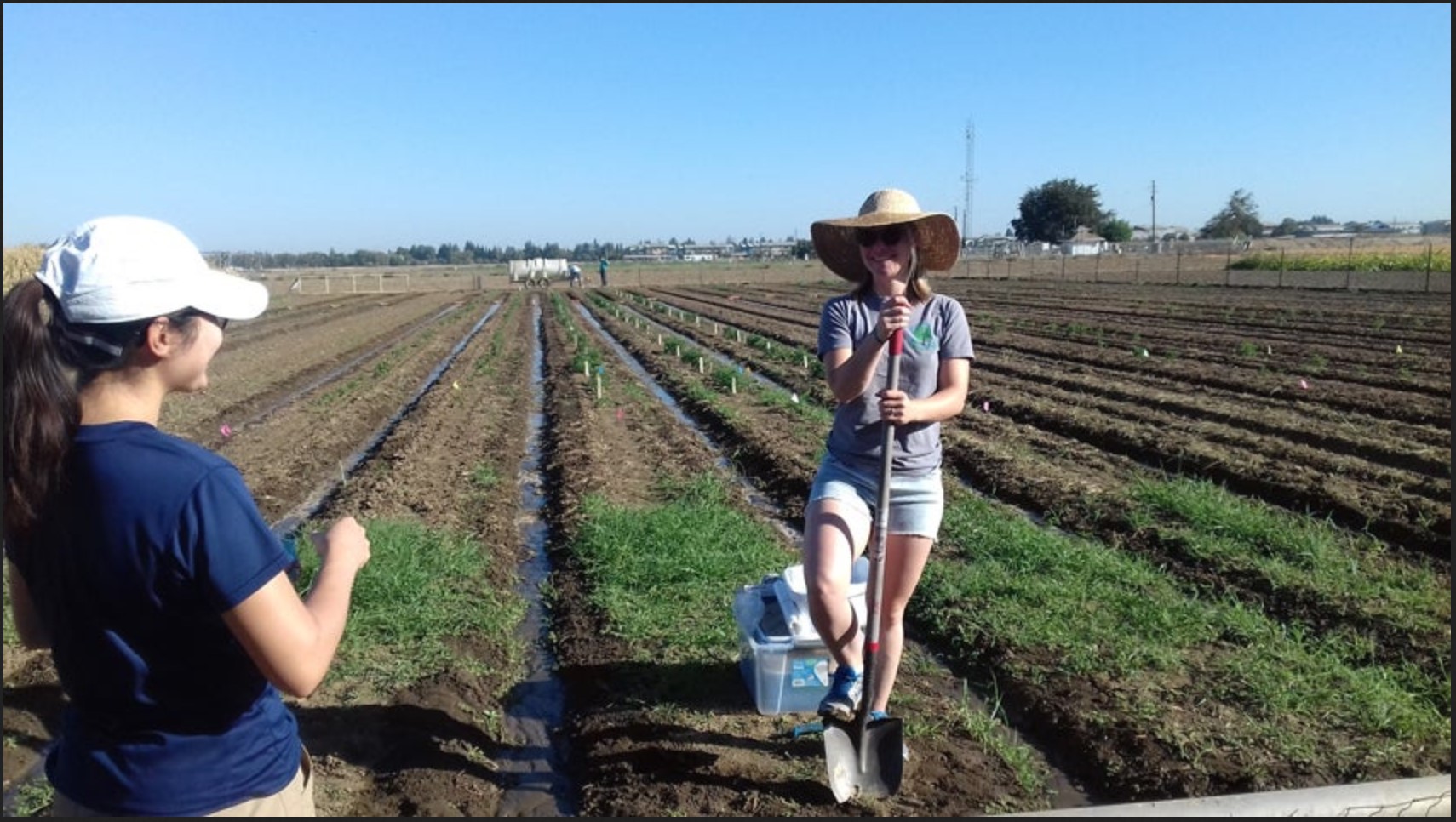 A woman standing in a plowed field with a wird-brimmed hat and shovel