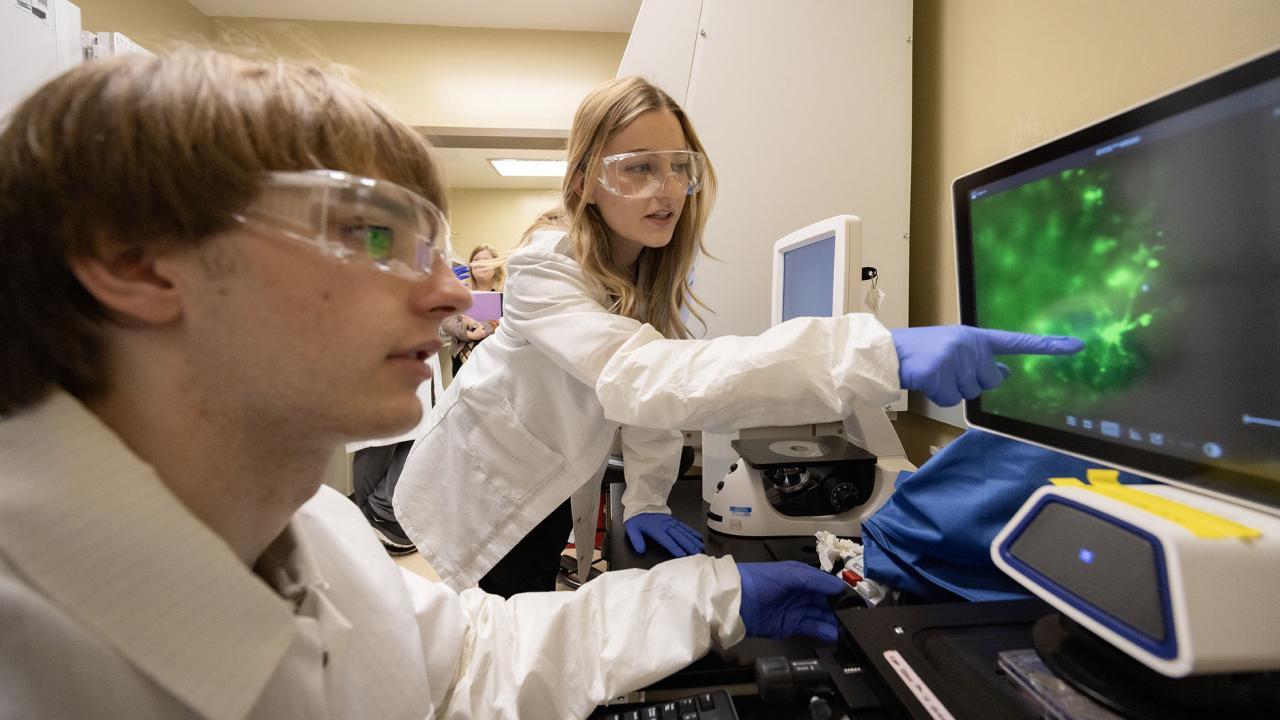Two students in lab coats and safety goggles examine green fluorescence on a computer screen.
