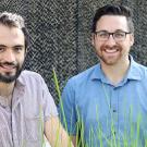 Two men smiling outdoors, standing among tall grass with a textured background.